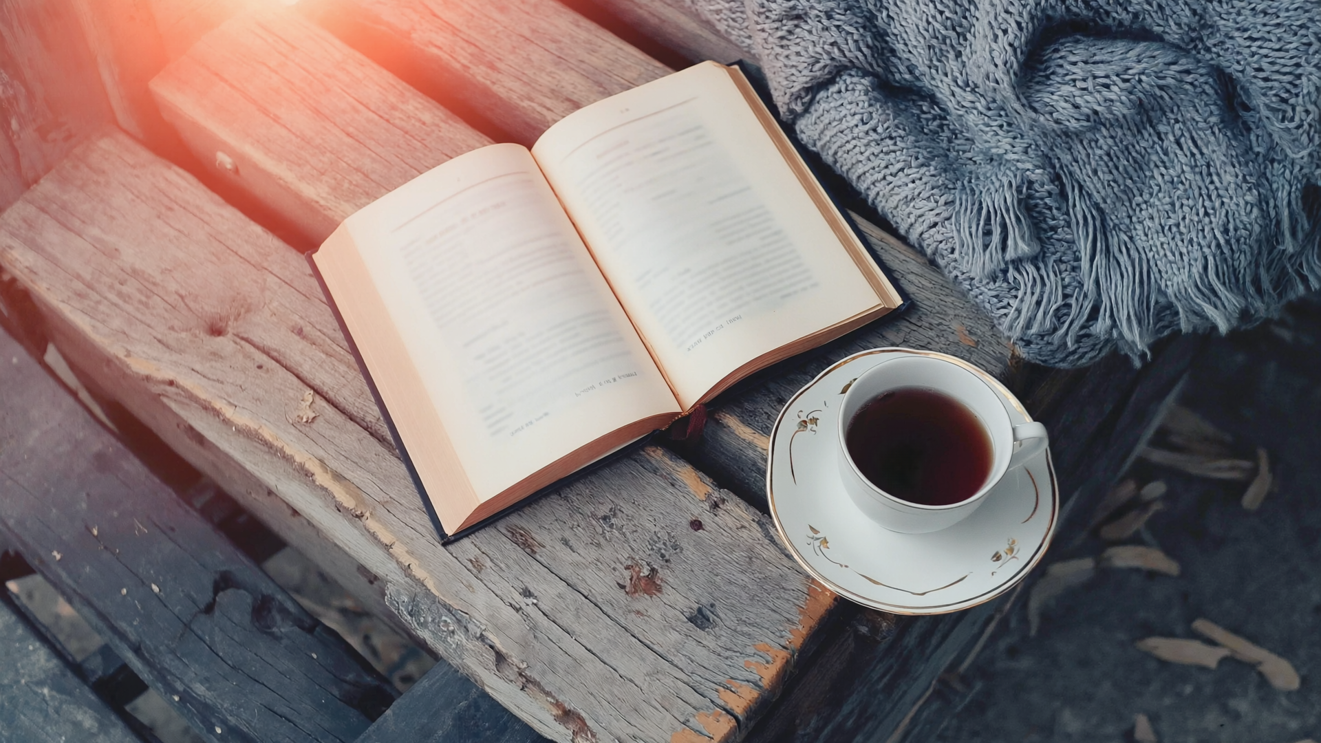 Aufgeschlagenes Sachbuch auf einer Holzbank neben einer Tasse Tee und einer gefalteten Wolldecke im weichen Nachmittagslicht.
